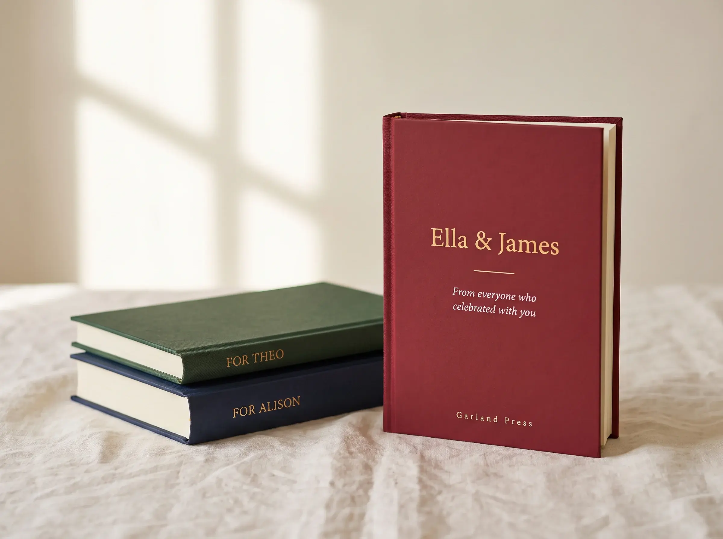 Three cloth-bound keepsake books: a standing burgundy book titled Ella & James in gold foil, with forest-green and navy books stacked beside it, their spines reading For Theo and For Peter